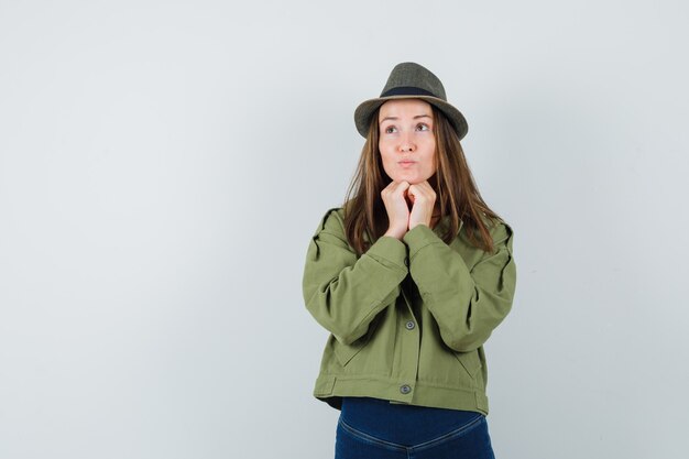 Junge Frau, die Kinn auf Händen in Jacke, Hose, Hut stützt und hoffnungsvoll aussieht. Vorderansicht.