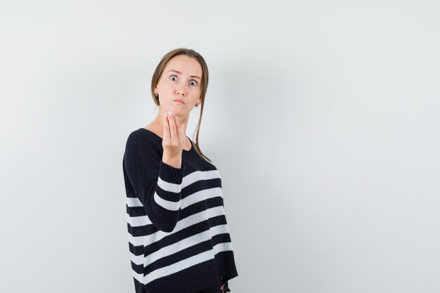 Junge Frau, die köstliche Geste in gestreiften Strickwaren und in schwarzen Hosen zeigt und ernst schaut