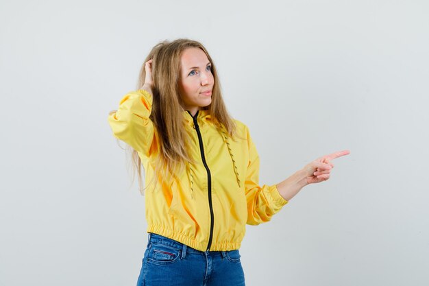 Junge Frau, die Kopf kratzt, während sie rechts in gelbe Bomberjacke und blaue Jeans zeigt und nachdenklich, Vorderansicht schaut.