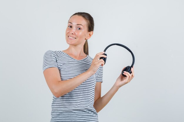 Junge Frau, die Kopfhörer im gestreiften T-Shirt hält und fröhlich schaut. Vorderansicht.