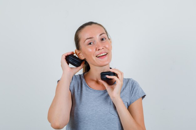 Junge Frau, die Kopfhörer nahe Ohr in der grauen T-Shirt-Vorderansicht hält.