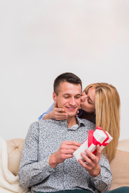 Junge Frau, die lächelnden Mann mit Geschenk auf Sofa küsst