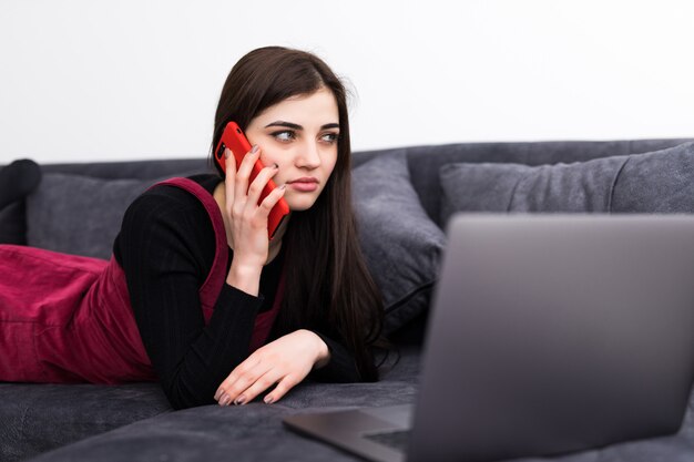 Junge Frau, die Laptop und Handy auf Sofa zu Hause verwendet