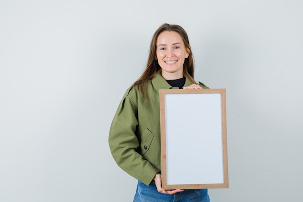 Junge Frau, die leeren Rahmen in grüner Jacke hält und zufrieden schaut. Vorderansicht.