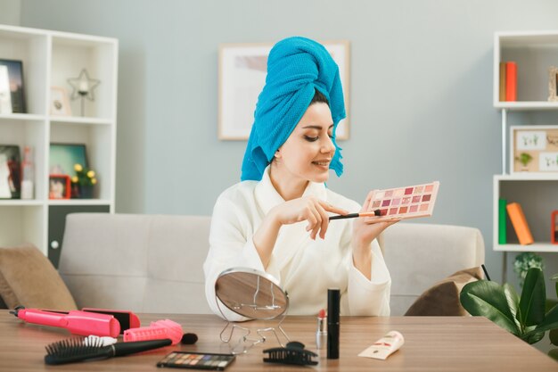 Junge Frau, die Lidschatten-Palette mit Make-up-Pinsel in Handtuch hält und betrachtet, sitzt am Tisch mit Make-up-Tools im Wohnzimmer