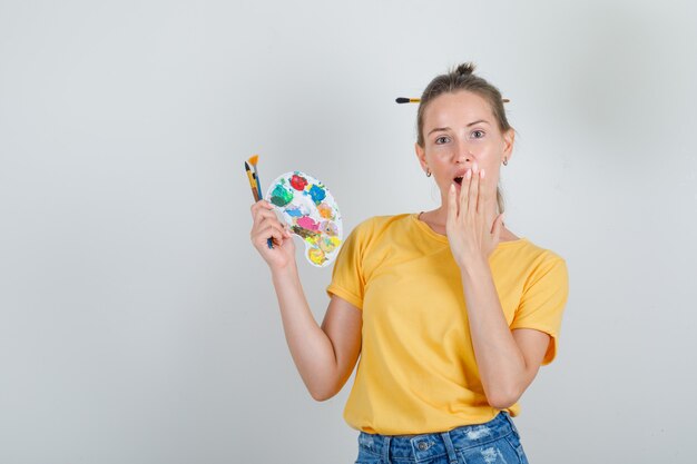 Junge Frau, die Malwerkzeuge mit Hand auf Mund in gelbem T-Shirt, Jeansshorts hält und überrascht schaut