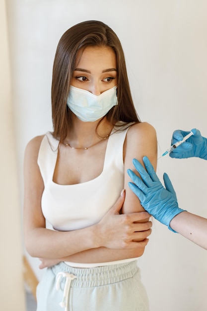 Junge Frau, die Maske trägt und Coronavirus-Impfstoff erhält Ein junges Mädchen, das einen Immunitätsschuss in den Unterarm erhält Nahaufnahme eines Arztes in blauen Handschuhen, der Covid19 AIDS gibt
