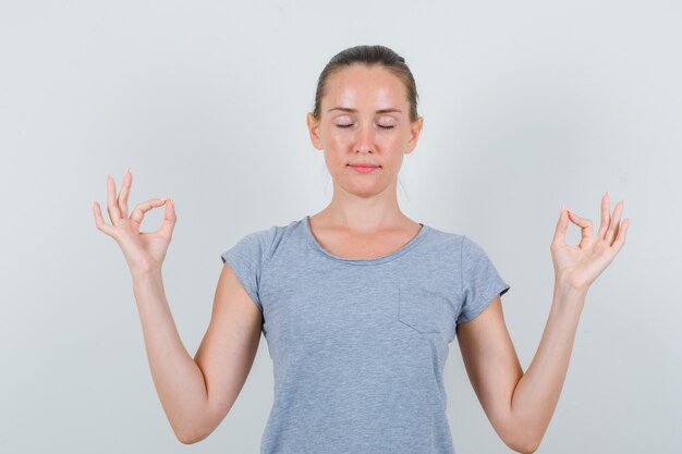 Junge Frau, die Meditation mit geschlossenen Augen im grauen T-Shirt tut und friedlich, Vorderansicht schaut.