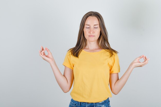 Junge Frau, die Meditation mit geschlossenen Augen im T-Shirt, in den Shorts tut und entspannt schaut