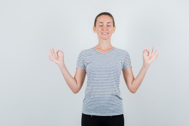 Junge Frau, die Meditation mit geschlossenen Augen im T-Shirt, in der Hose tut und entspannt schaut. Vorderansicht.