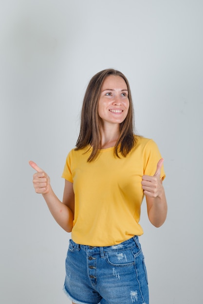 Junge Frau, die mit Daumen hoch im gelben T-Shirt, in den Shorts weg schaut und fröhlich schaut