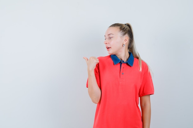 Junge Frau, die mit Daumen im T-Shirt beiseite zeigt und konzentriert schaut. Vorderansicht.