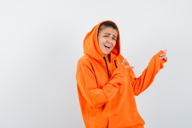 Kostenloses Foto junge frau, die mit den zeigefingern im orangefarbenen hoodie nach rechts zeigt und glücklich aussieht