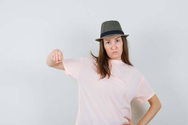 Junge Frau, die mit Faust in rosa T-Shirt, Hut droht und ernst schaut. Vorderansicht.