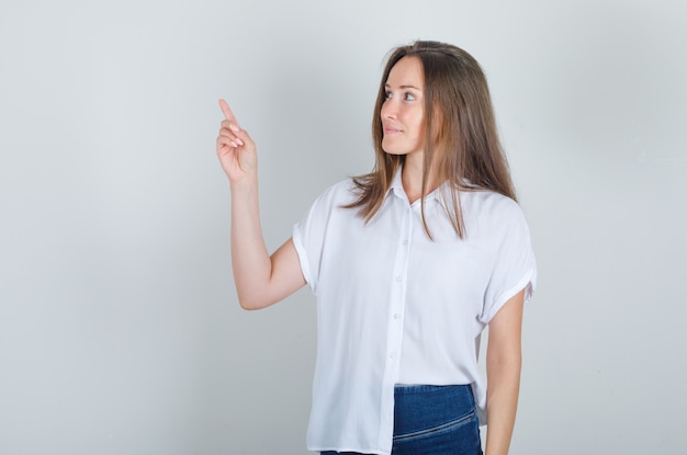 Junge Frau, die mit Finger oben im weißen T-Shirt, in Jeans wegschaut und froh schaut