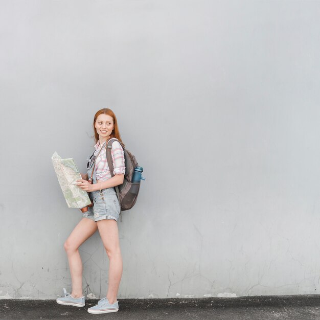 Junge Frau, die mit Karte und Rucksack nahe Wand steht