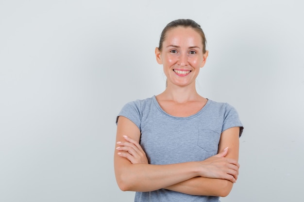 Junge Frau, die mit verschränkten Armen im grauen T-Shirt steht und fröhlich schaut. Vorderansicht.