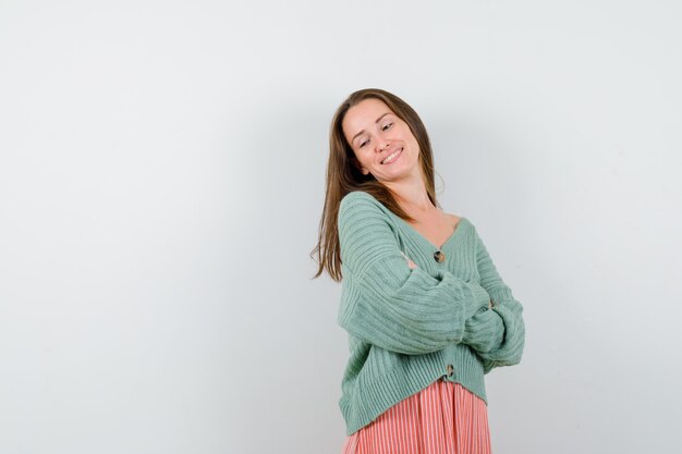 Junge Frau, die mit verschränkten Armen in Strickjacke, Rock steht und fröhlich schaut, Vorderansicht.