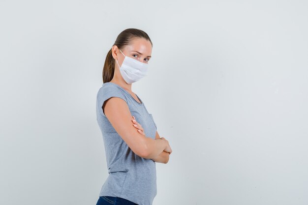 Junge Frau, die mit verschränkten Armen in T-Shirt, Maske, Jeans steht und fröhlich schaut.