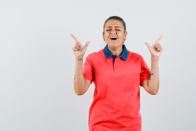 Junge Frau, die mit Zeigefingern im roten T-Shirt nach oben zeigt und amüsiert aussieht. Vorderansicht.