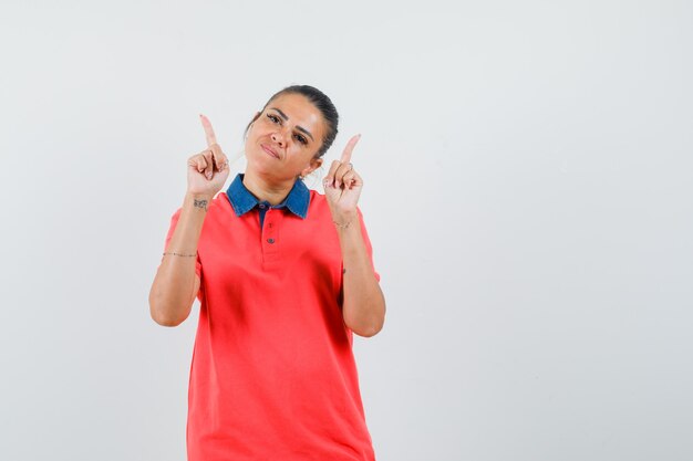 Junge Frau, die mit Zeigefingern im roten T-Shirt oben zeigt und hübsch, Vorderansicht schaut.