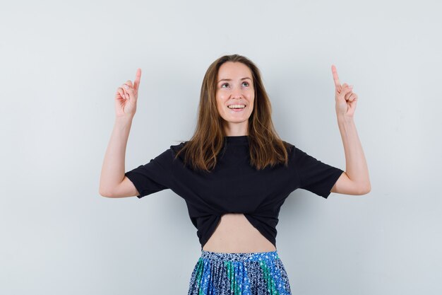 Junge Frau, die mit Zeigefingern im schwarzen T-Shirt und im blauen Rock oben zeigt und glücklich schaut