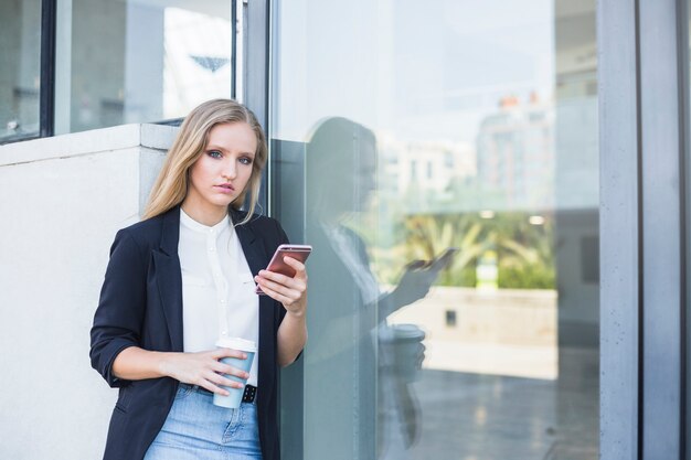 Junge Frau, die MitnehmerKaffeetasse und Handy sich lehnt auf reflektierendem Glas hält