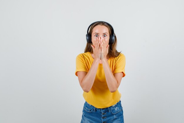 Junge Frau, die Mund mit Händen im T-Shirt, in den Shorts, in den Kopfhörern bedeckt und ängstlich schaut