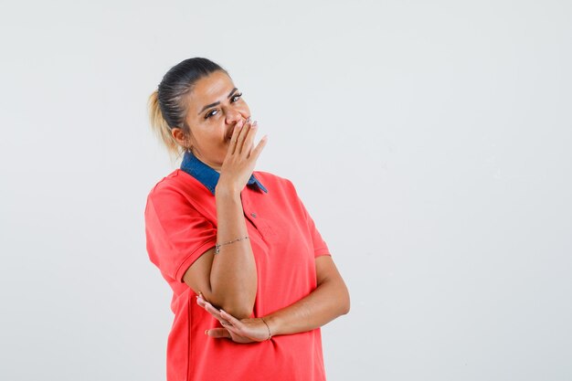 Junge Frau, die Mund mit Hand im roten T-Shirt bedeckt und glücklich schaut. Vorderansicht.