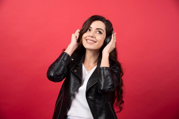 Junge Frau, die Musik über Kopfhörer hört und auf einem roten Hintergrund aufwirft. Foto in hoher Qualität