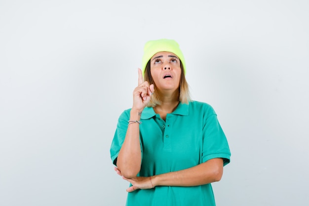 Junge Frau, die nach oben zeigt, im Polo-T-Shirt, in der Mütze nach oben schaut und nachdenklich aussieht. Vorderansicht.