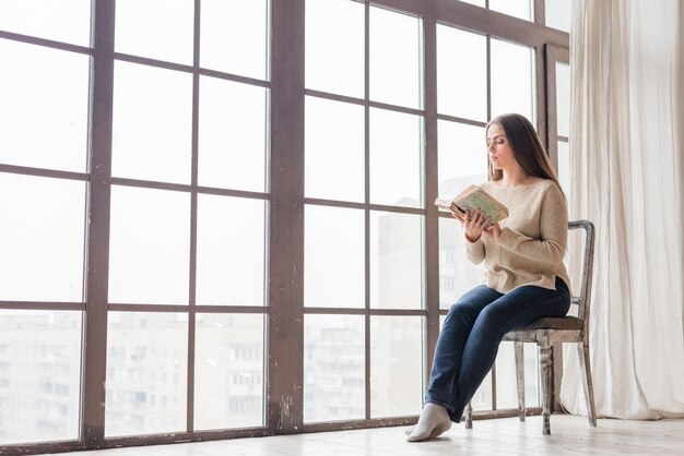 Junge Frau, die nahe dem Fenster liest das Buch sitzt