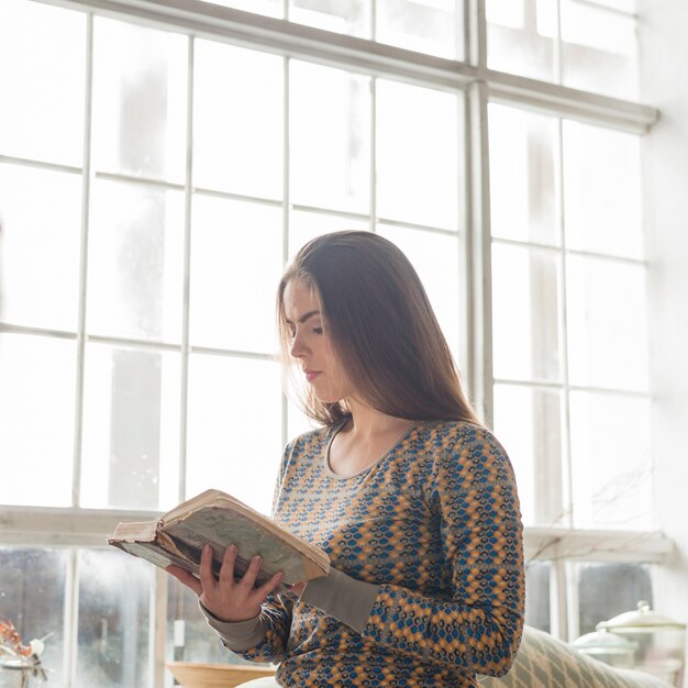 Junge Frau, die nahe dem Fenster steht und ein altes Buch liest