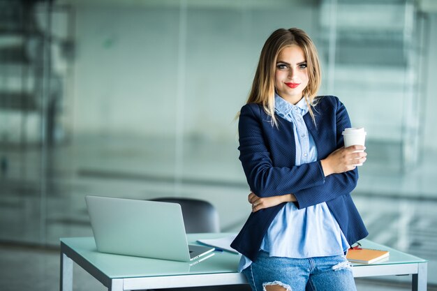 Junge Frau, die nahe Schreibtisch mit Laptop hält Ordner und Tasse Kaffee steht. Arbeitsplatz. Geschäftsfrau.