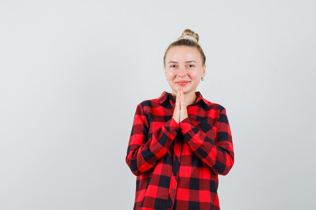 Junge Frau, die namaste Geste im karierten Hemd zeigt und fröhlich, Vorderansicht schaut.