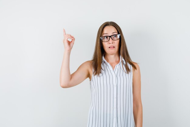 Junge Frau, die oben im T-Shirt zeigt und verwirrt, Vorderansicht schaut.