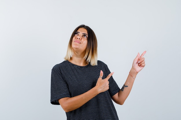 Junge Frau, die obere rechte Seite mit Zeigefingern im schwarzen Kleid zeigt und konzentriert schaut. Vorderansicht.