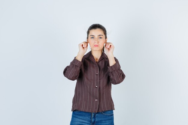 Junge Frau, die Ohrläppchen in gestreiftem Hemd, Jeans zerrt und süß aussieht. Vorderansicht.