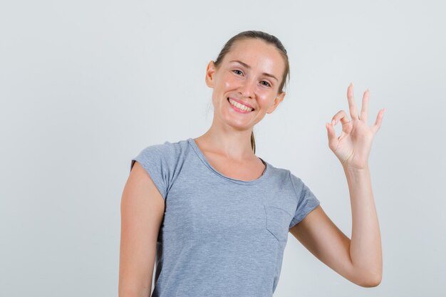 Junge Frau, die ok Geste im grauen T-Shirt zeigt und erfreut schaut. Vorderansicht.