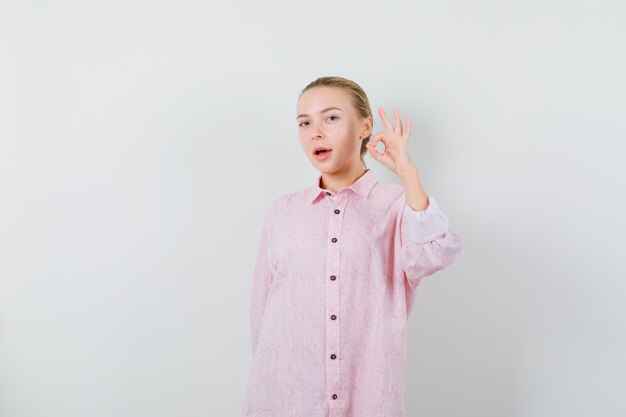 Junge Frau, die ok Geste im rosa Hemd zeigt und erfreut schaut