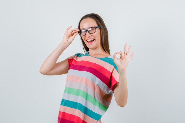 Junge Frau, die ok Geste mit den Fingern auf Brille im T-Shirt zeigt und fröhlich schaut. Vorderansicht.
