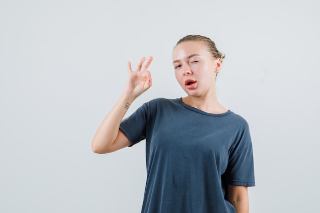 Junge Frau, die ok Geste und zwinkerndes Auge im grauen T-Shirt zeigt