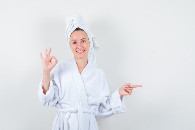 Junge Frau, die ok Geste zeigt, rechts im weißen Bademantel, Handtuch zeigt und fröhlich schaut, Vorderansicht.