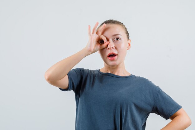 Junge Frau, die ok Zeichen auf Auge im grauen T-Shirt zeigt und erstaunt schaut