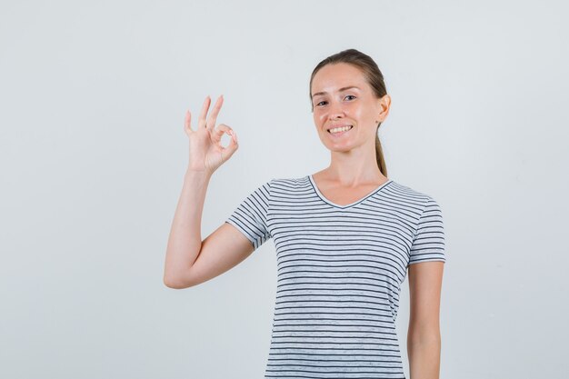 Junge Frau, die ok Zeichen im gestreiften T-Shirt zeigt und fröhlich schaut. Vorderansicht.