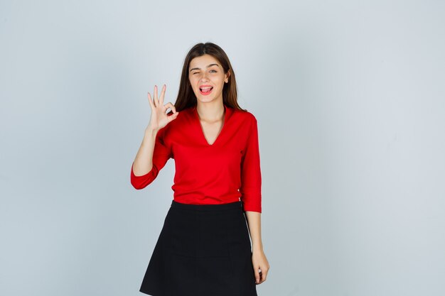Junge Frau, die ok Zeichen zeigt, Zunge heraus in rote Bluse stehend