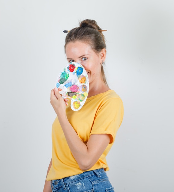 Junge Frau, die Palette über Gesicht in gelbem T-Shirt, Jeansshorts hält und fröhlich schaut