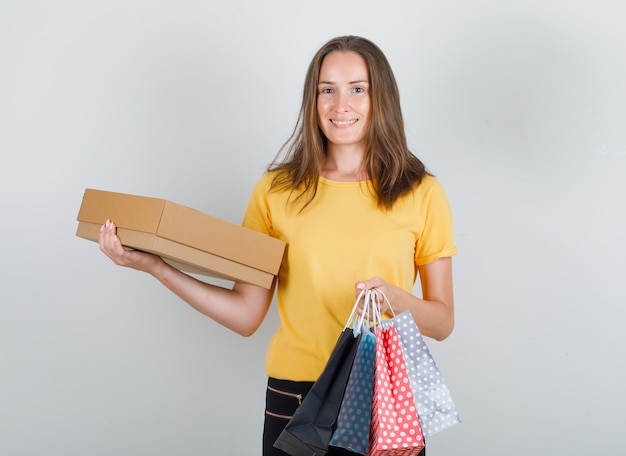 Junge Frau, die Papiertüten und Pappkarton im gelben T-Shirt, in den Hosen hält und froh schaut