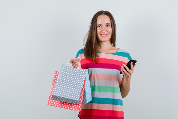 Junge Frau, die Papiertüten und Smartphone im T-Shirt hält und fröhlich schaut. Vorderansicht.