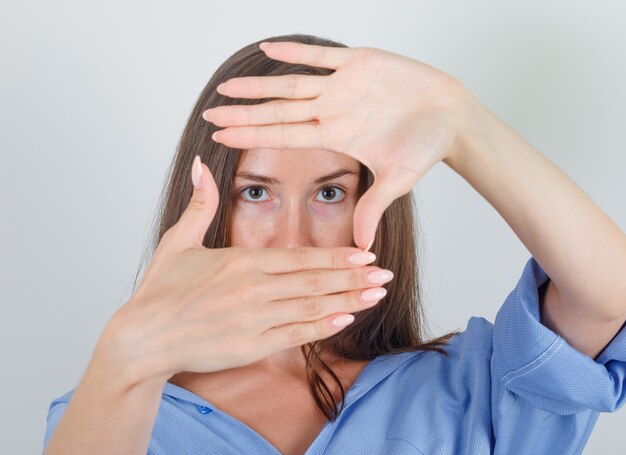 Junge Frau, die Rahmen der Finger im blauen Hemd macht und zuversichtlich schaut